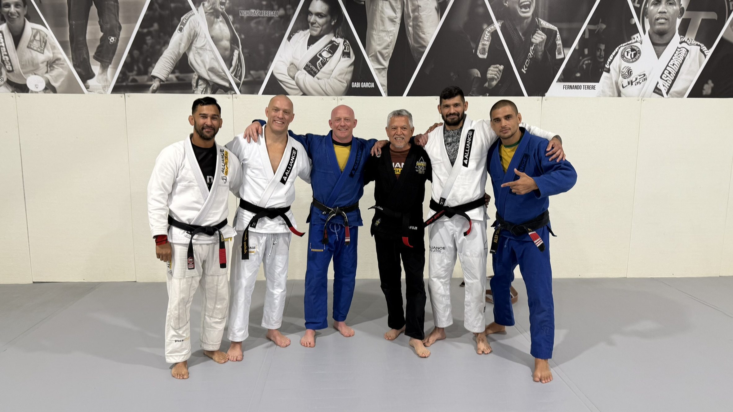 Six Alliance BJJ Houston coaches standing together in front of the founders' photo wall