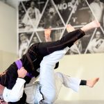 White-gi grappler sweeping a purple belt training partner during a live BJJ round