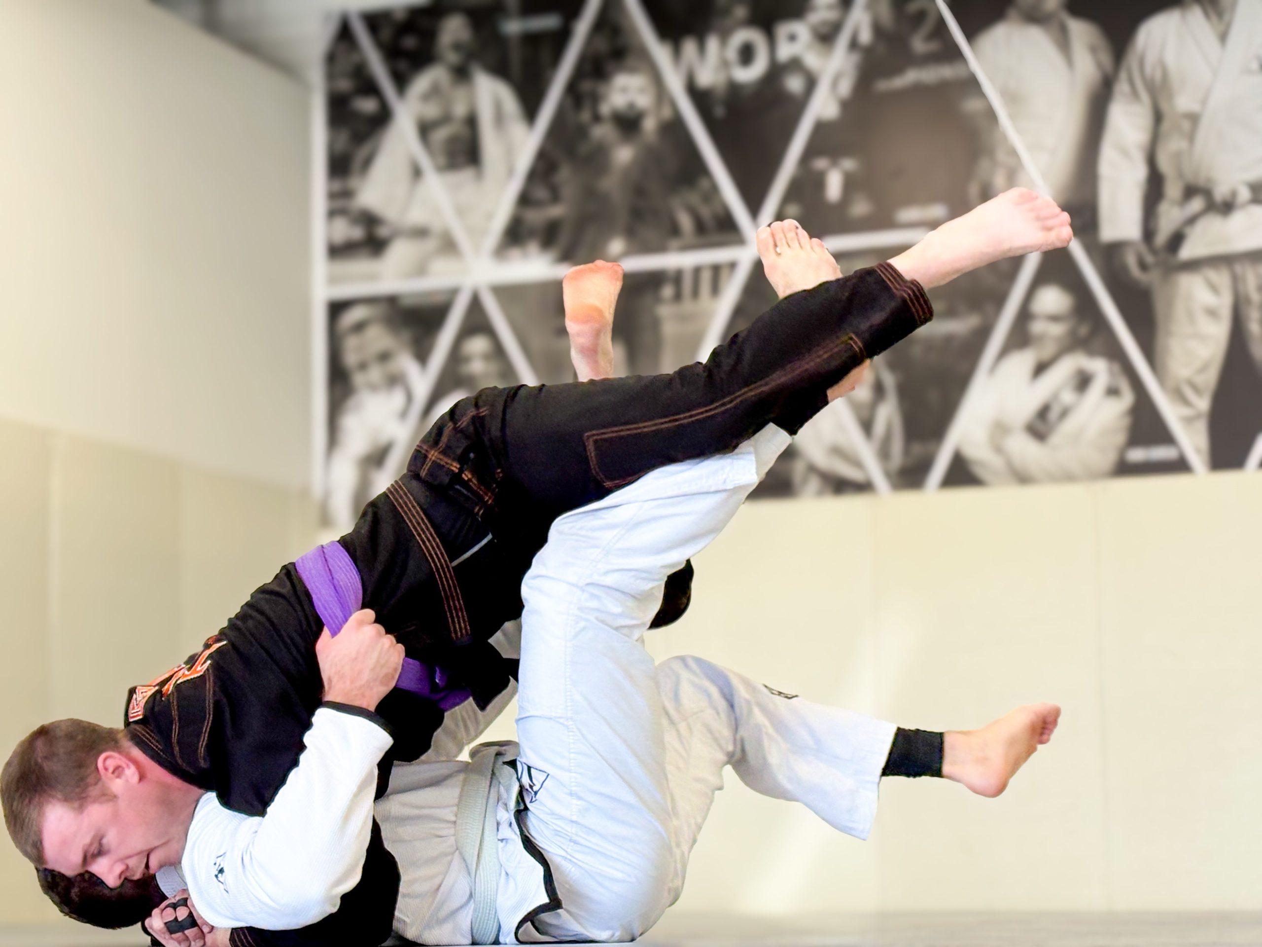 White-gi grappler sweeping a purple belt training partner during a live BJJ round