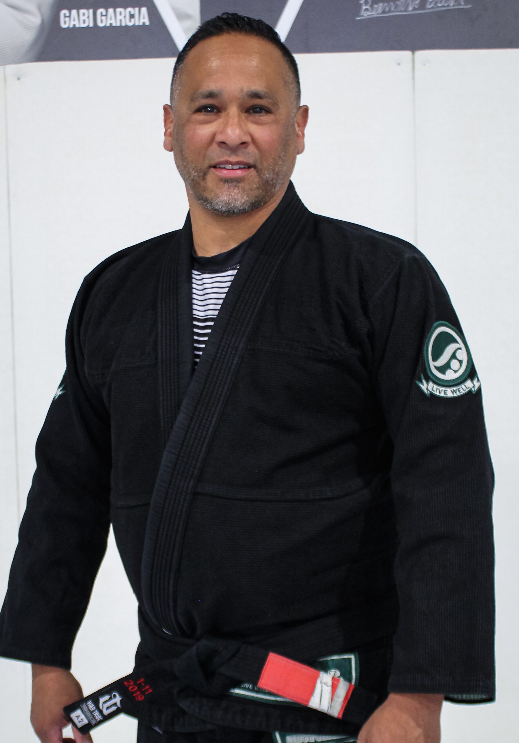 Coach John in a black Alliance kimono in front of the academy photo wall