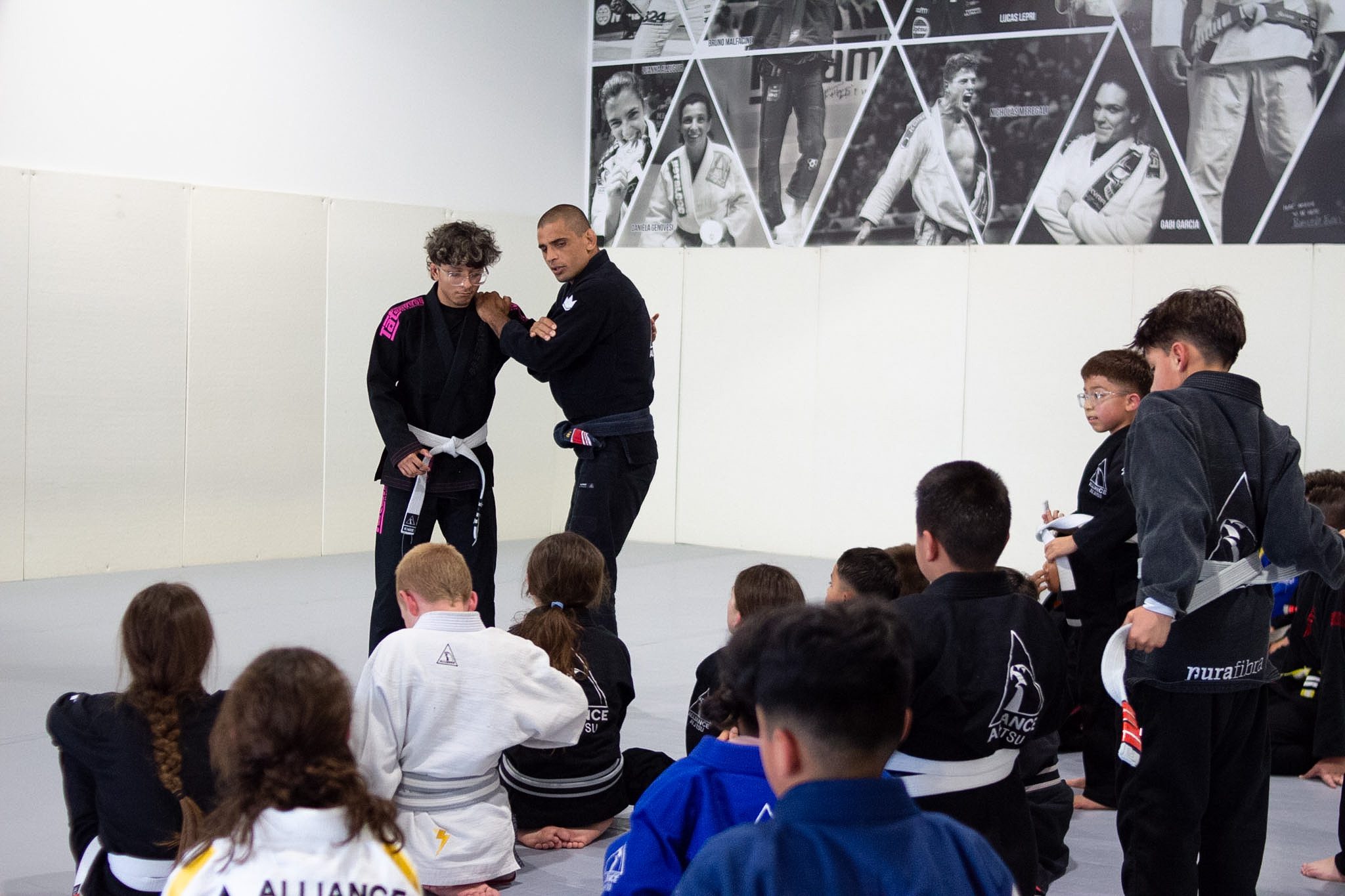 Coach showing a standing technique while kids watch from the mat