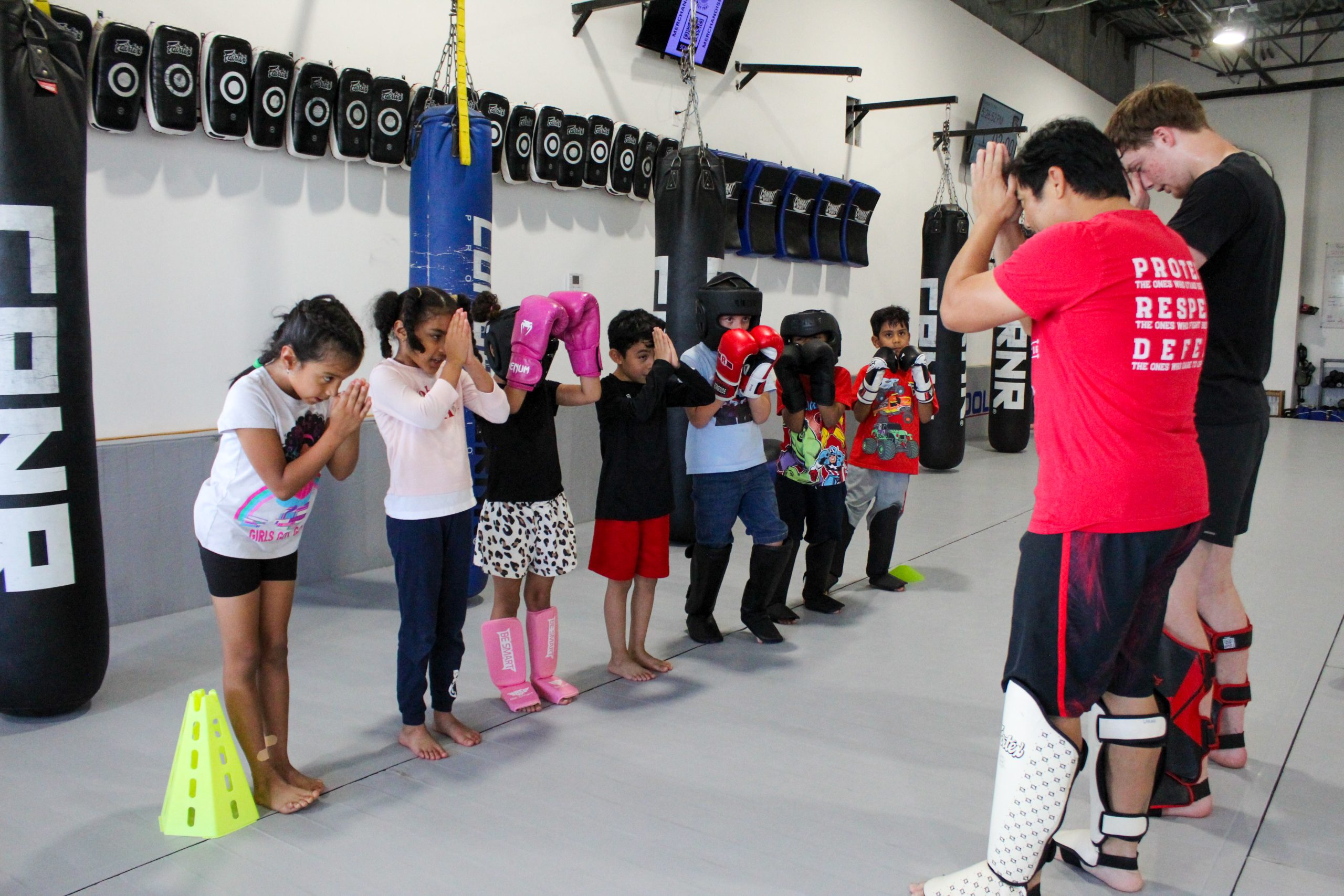 Kids in shin guards practicing Muay Thai guard position with a coach beside heavy bags