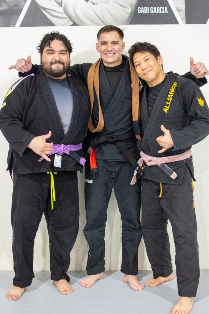 Thomas Dougherty in a black gi and brown belt sitting on the mat smiling