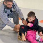 Coach in a gray gi guiding two preschool-age kids through a beginner BJJ drill