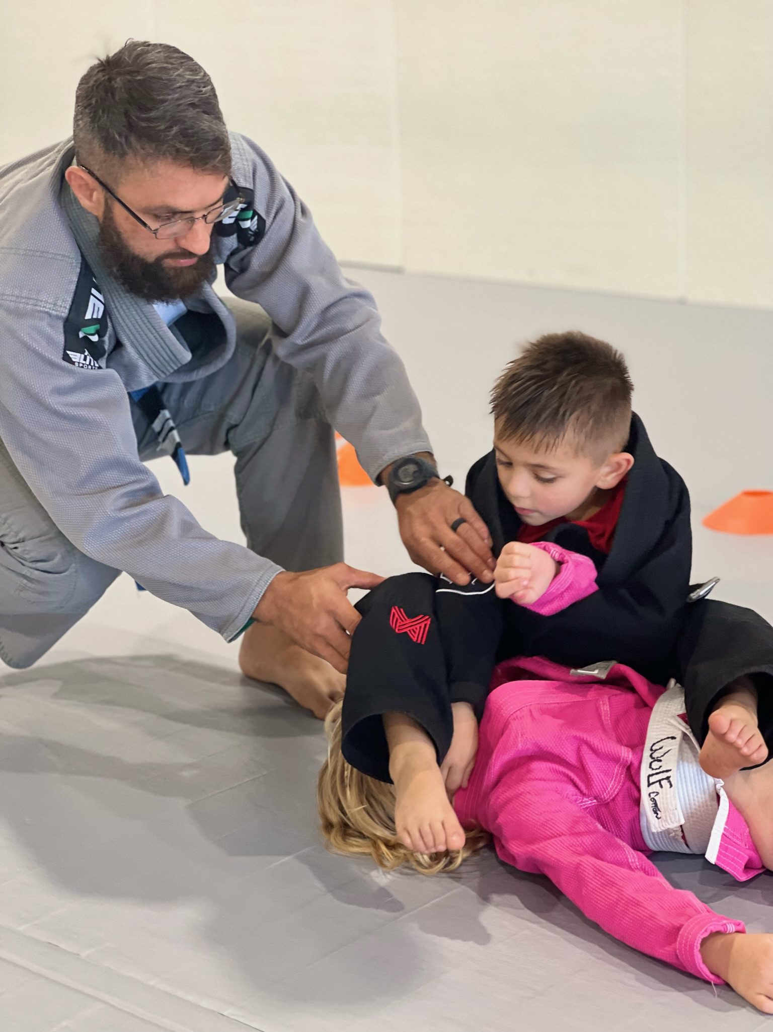 Coach in a gray gi guiding two preschool-age kids through a beginner BJJ drill