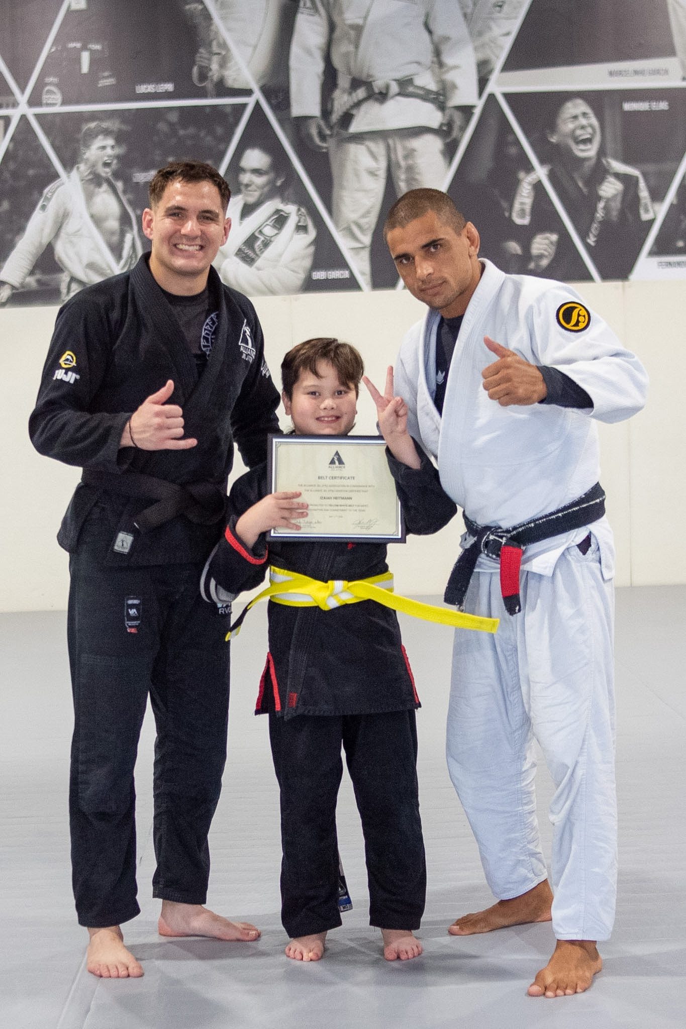 Junior Eagle in black gi holds belt certificate between two Alliance coaches