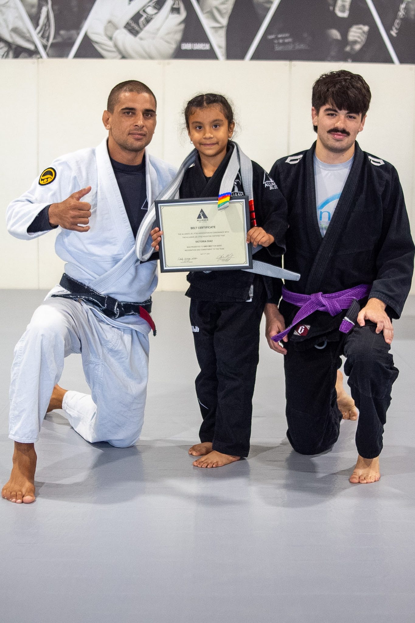 Young girl in black gi holds certificate between two Alliance jiu-jitsu coaches