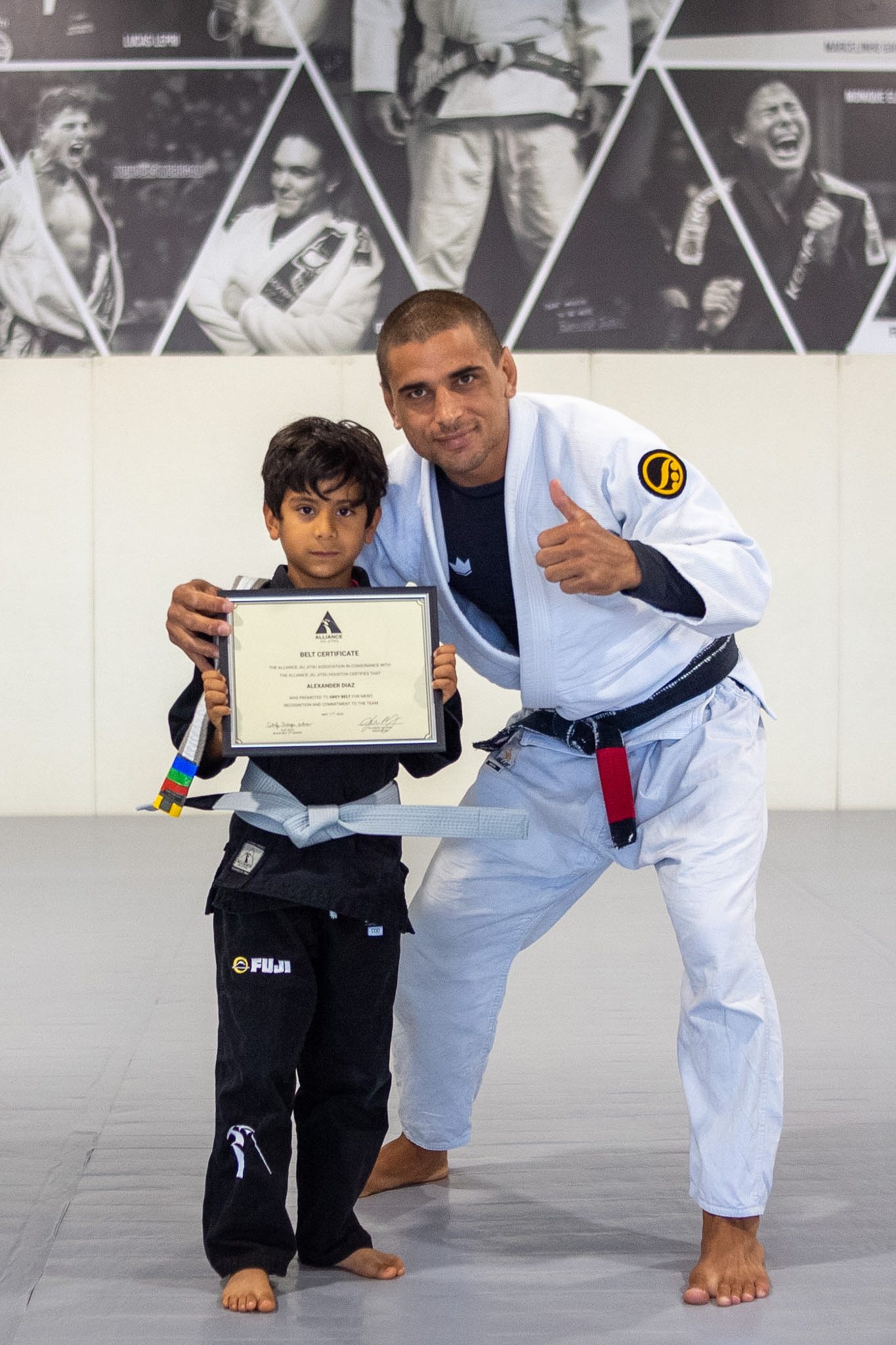 Little Eagle student in black gi holds Alliance belt certificate with head instructor