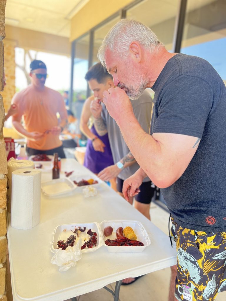 Bearded member tasting fresh boiled crawfish at outdoor tables under shaded patio