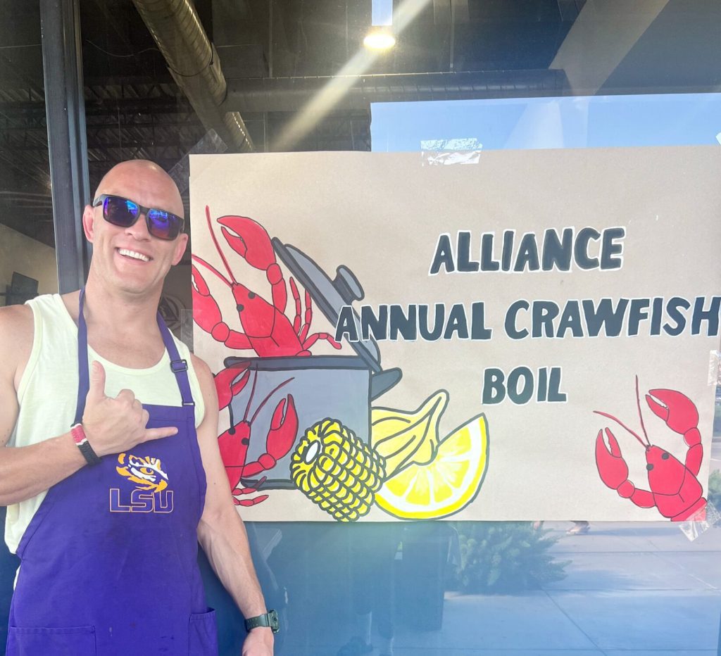 Tony Landry in LSU shirt and purple apron next to hand-painted crawfish boil banner