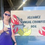 Tony Landry in LSU shirt and purple apron next to hand-painted crawfish boil banner