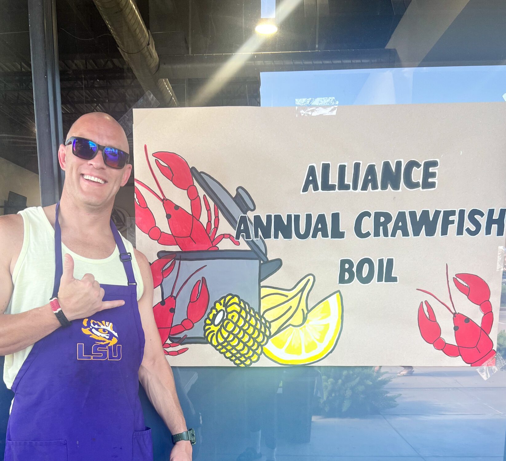 Tony Landry in LSU shirt and purple apron next to hand-painted crawfish boil banner