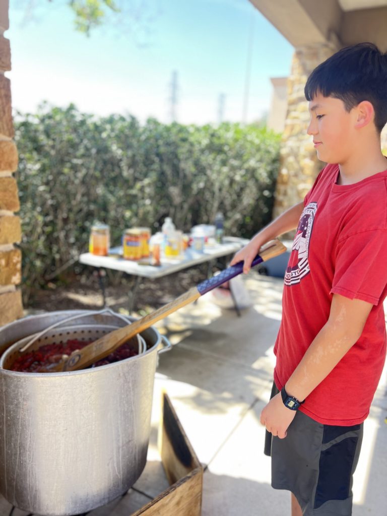 Boy in red shirt stirring large outdoor crawfish boil pot with long wooden paddle