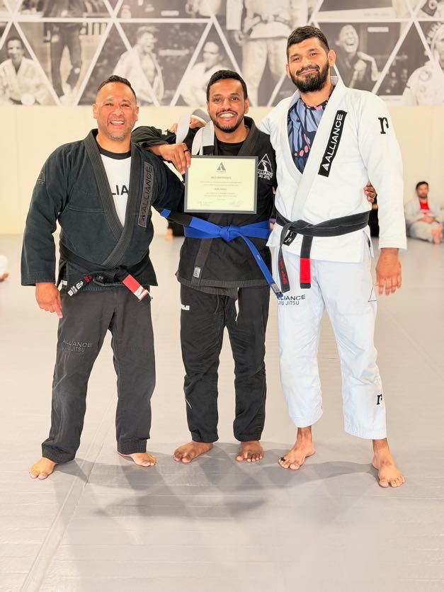 Smiling man in a black gi holds his certificate between two Alliance coaches