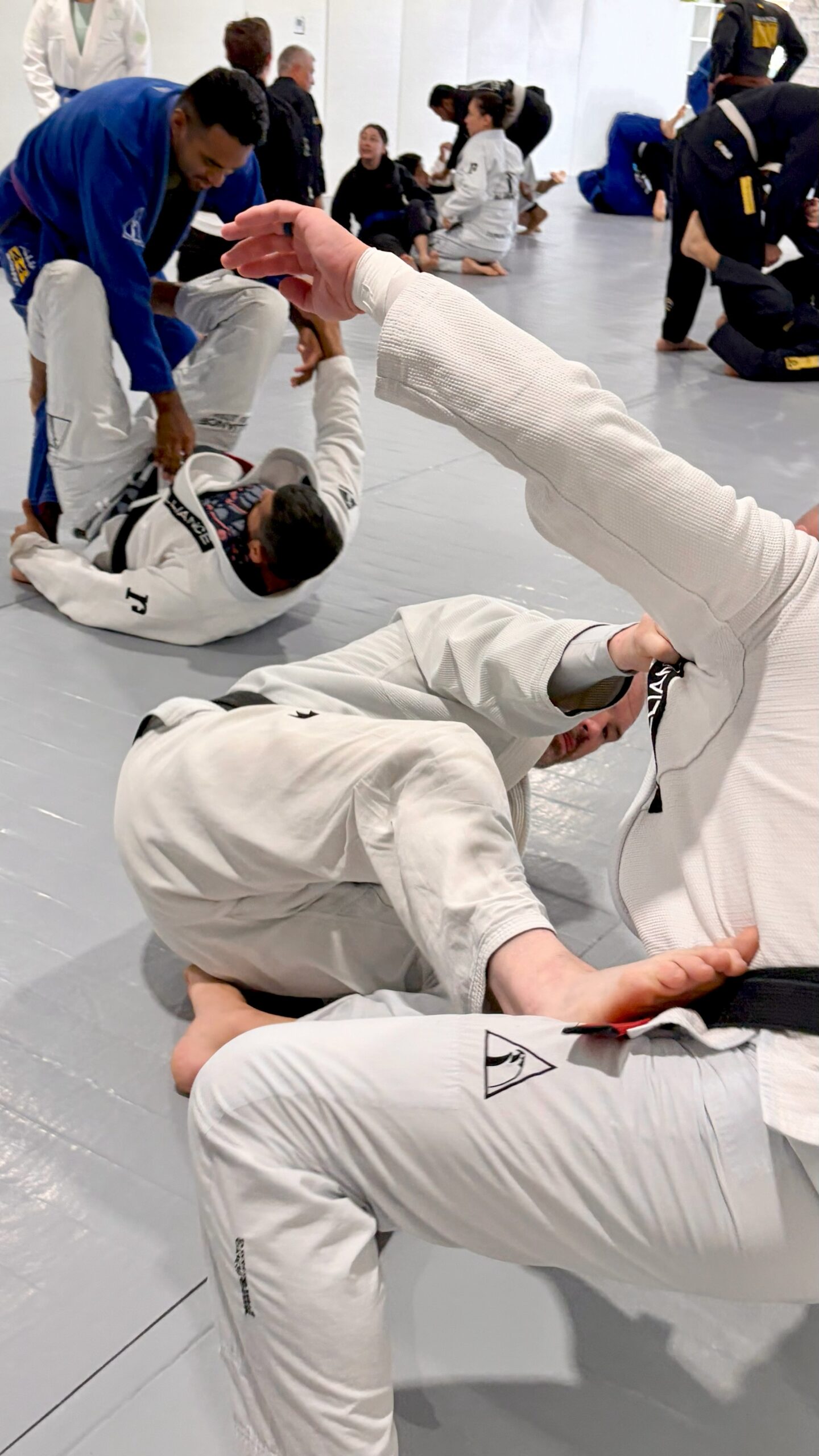 Two men in white gis grapple closely during a busy adult open mat session