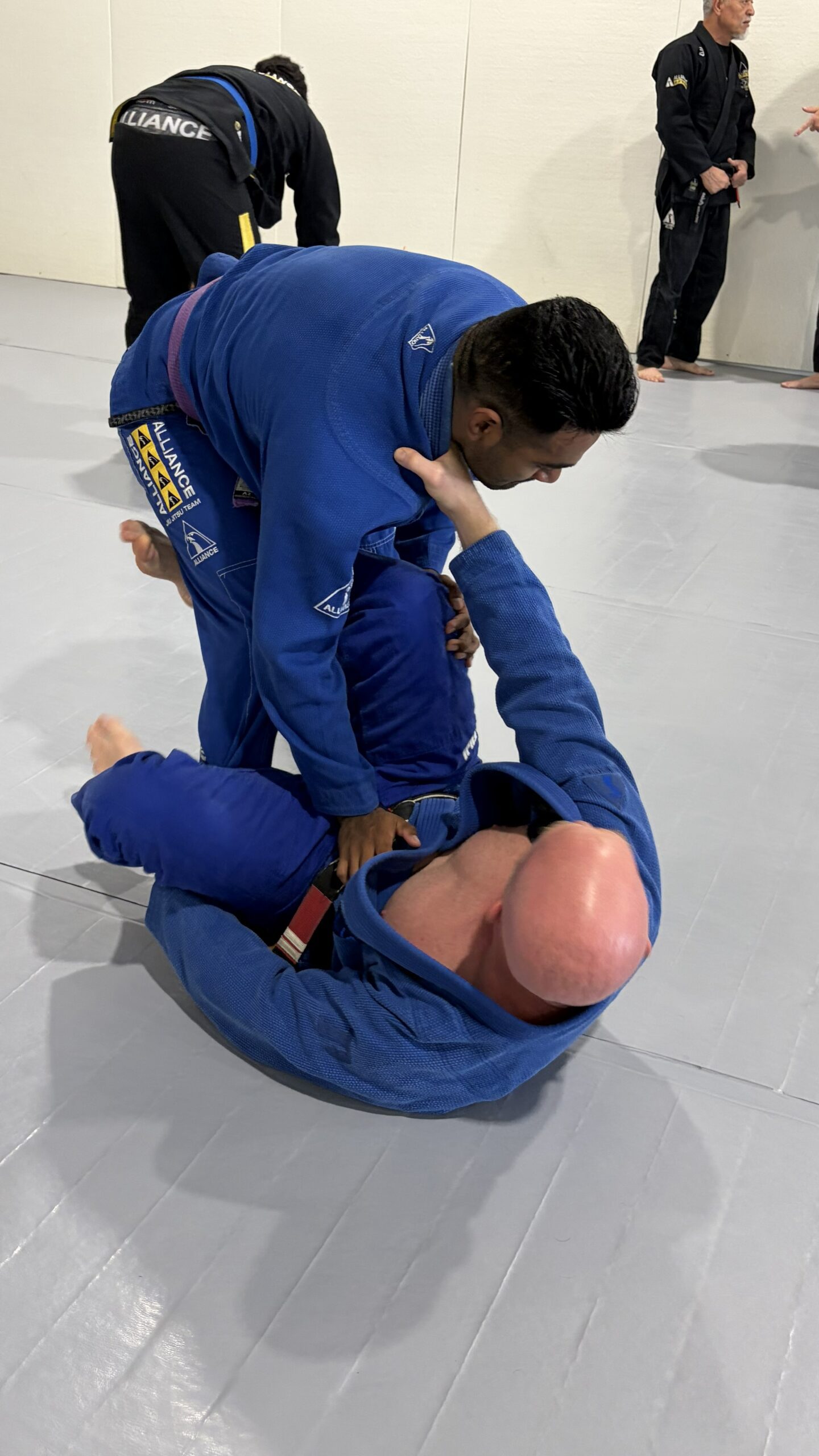 Man in a blue gi passes a partner's guard as a brown belt watches