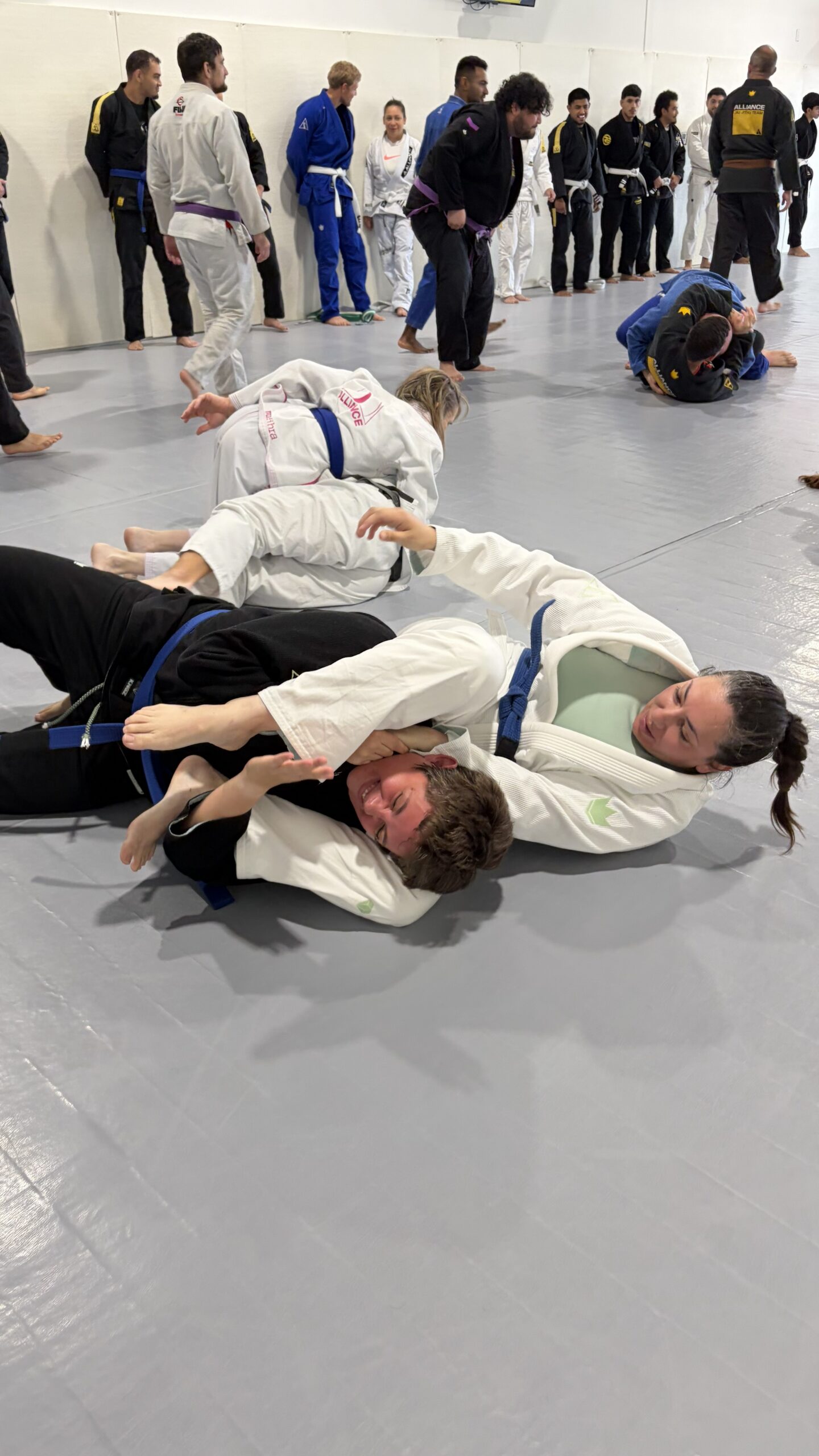 Two women grapple on the ground as a long row of teammates watches from the wall