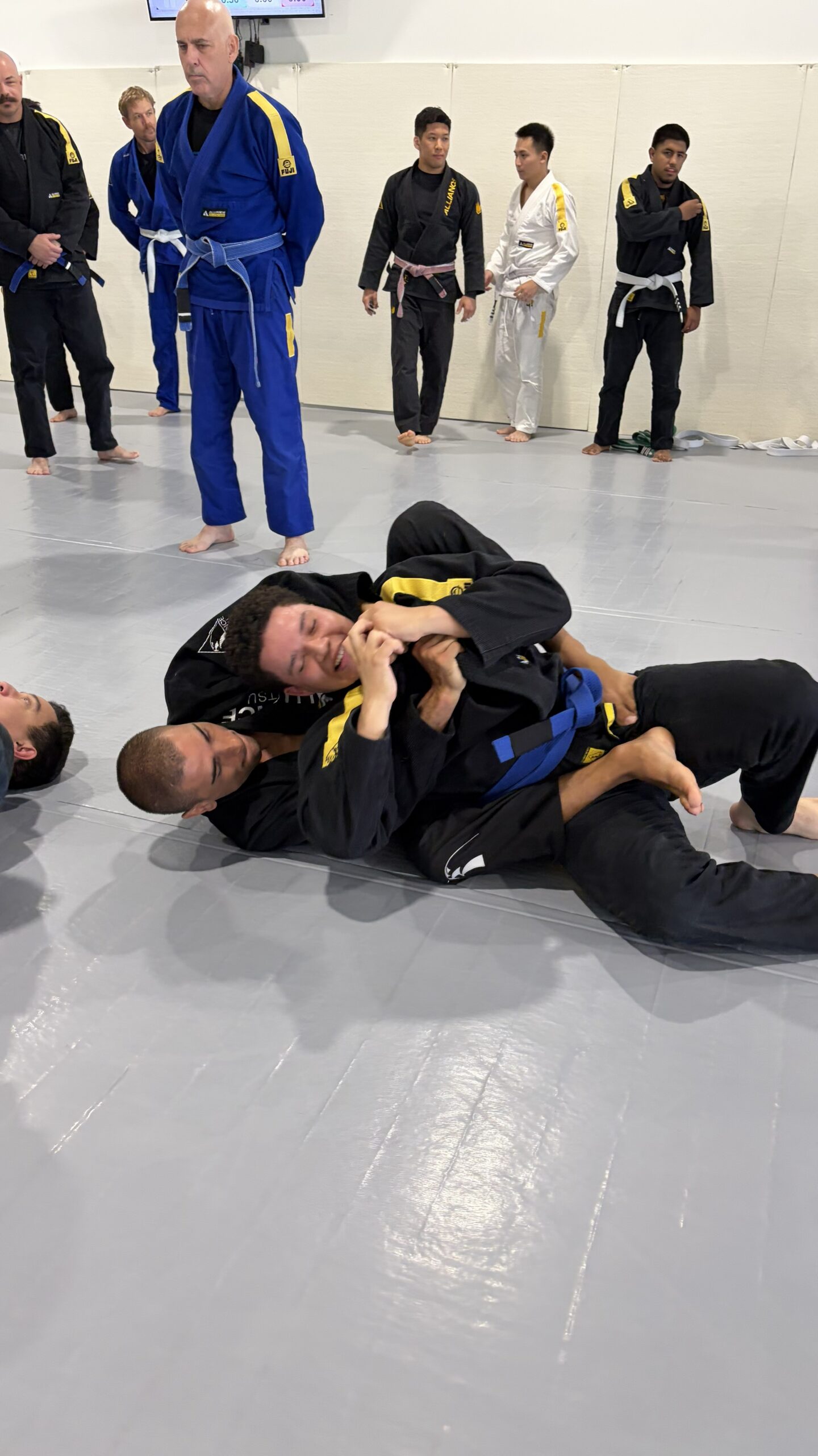 Two men in black gis grapple on the ground as teammates watch from the wall