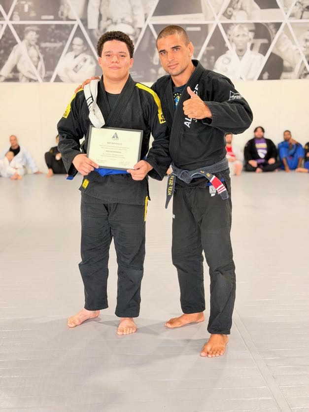 Man in a black gi holds his certificate and new blue belt beside a coach