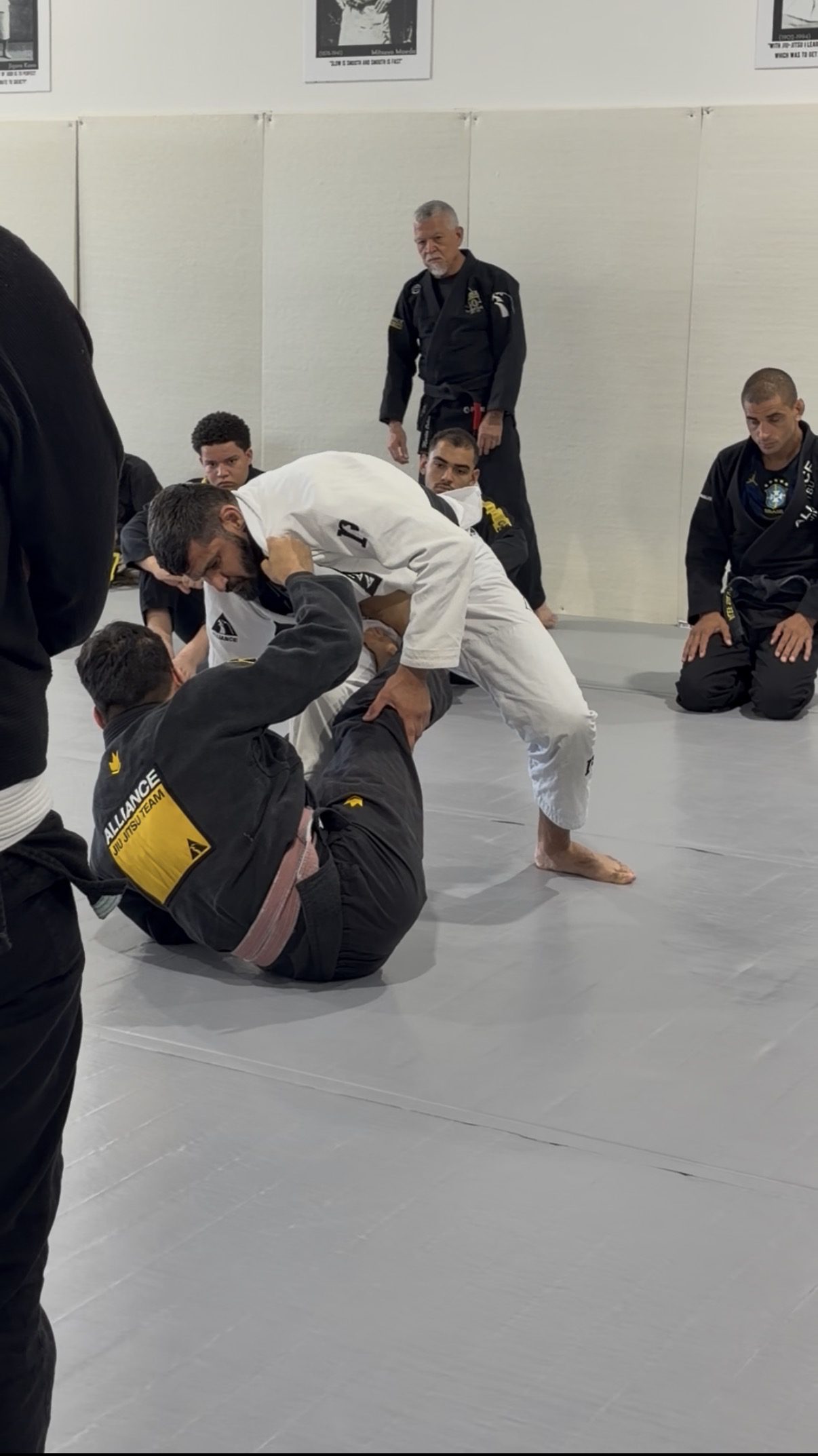 Man in a white gi drills a standing clinch as a coach watches nearby