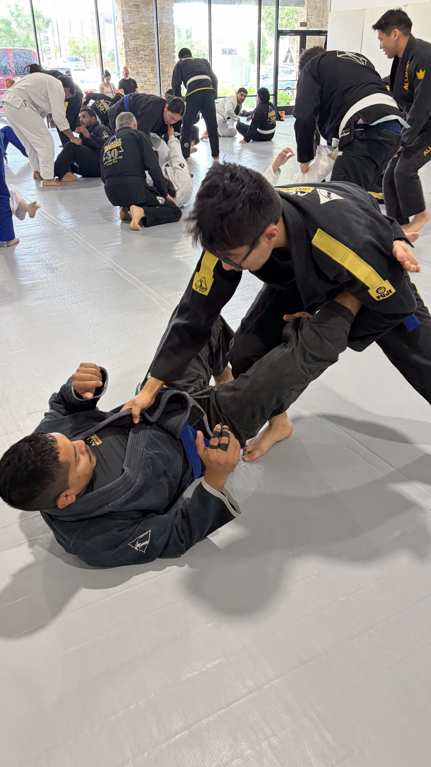Teen in a black gi and black belt controls a grounded partner during busy open mat