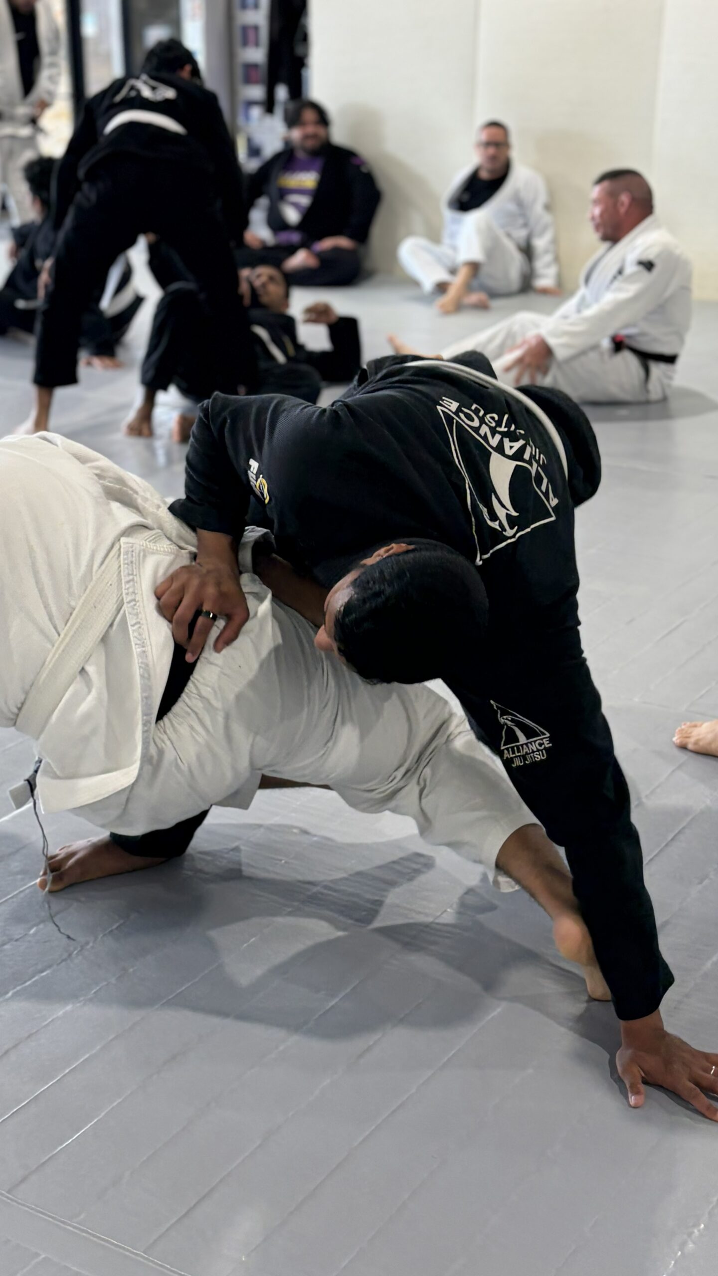 Man in a black Alliance gi drives a takedown on a partner in a white gi