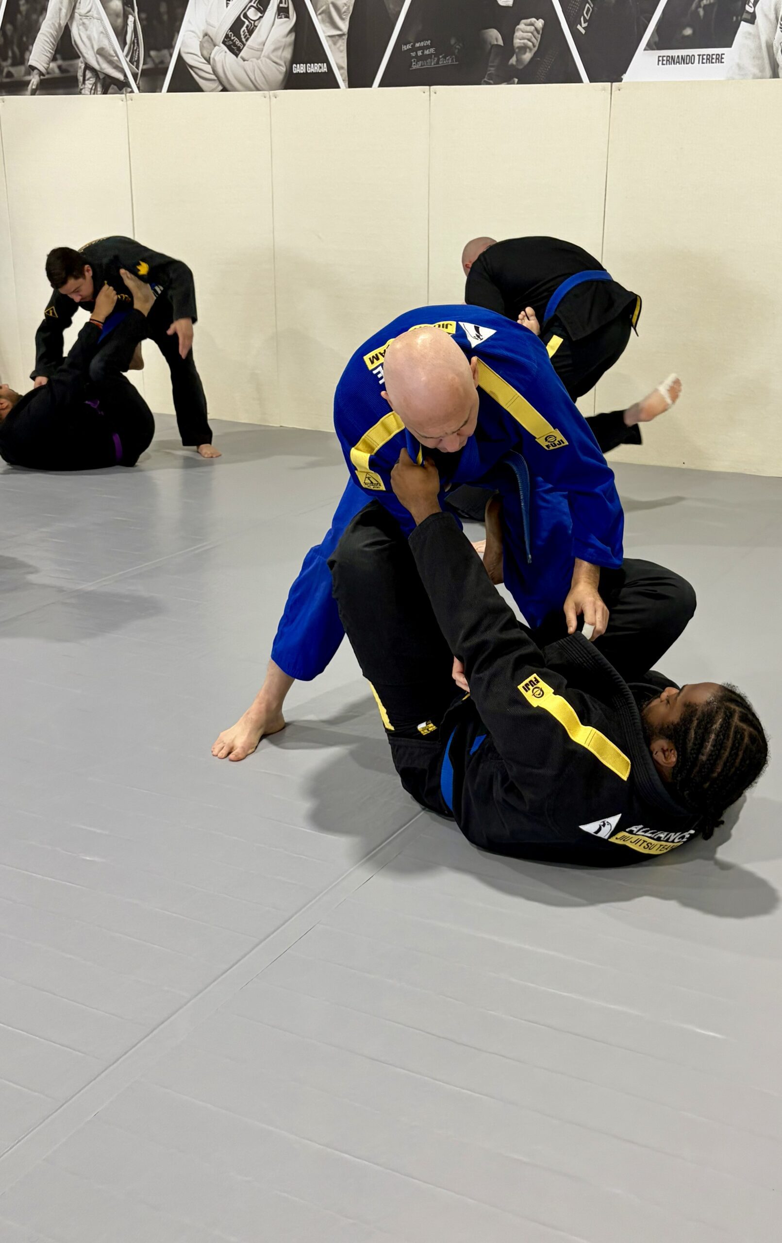 Man in a blue gi grips a downed partner during open mat sparring