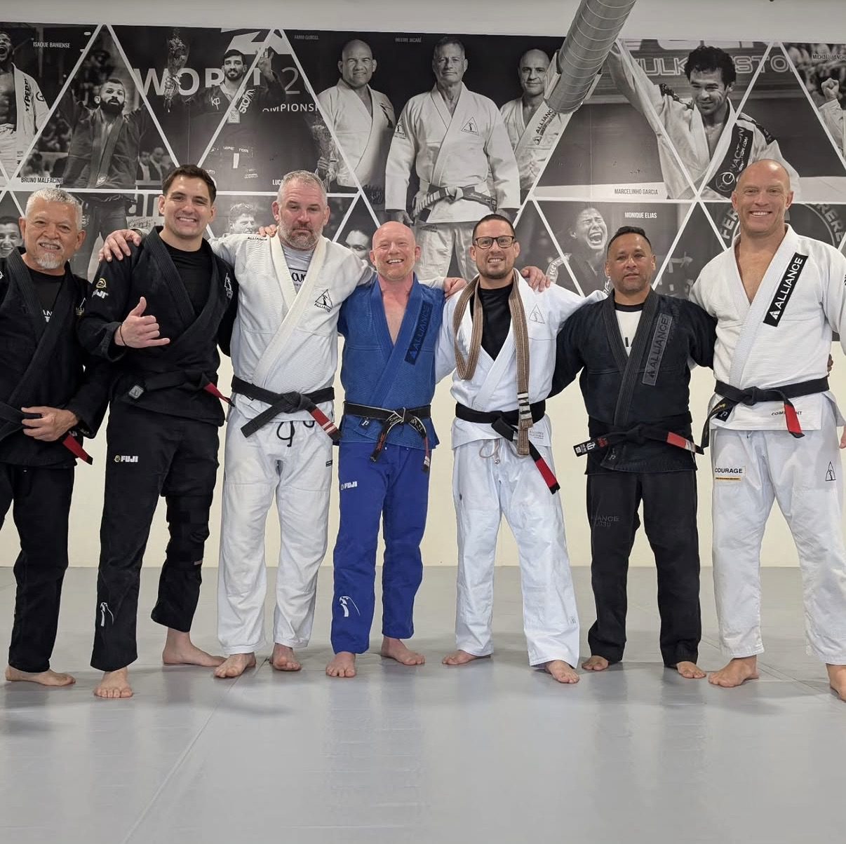 Seven adult men in mixed gis line up arm-in-arm wearing colored and black belts