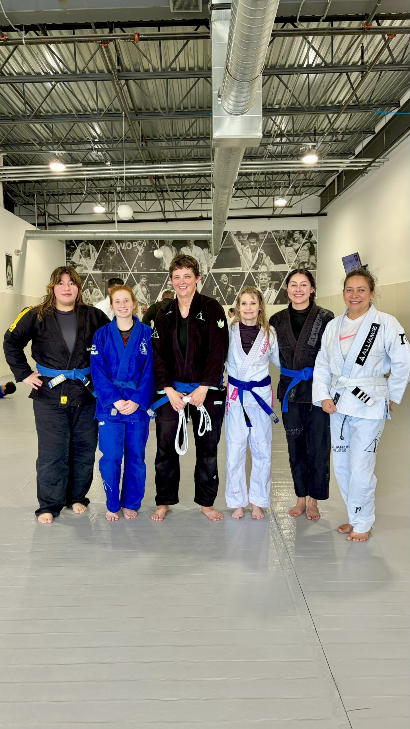 Alliance women grapplers pose together with new belts by the academy wall mural