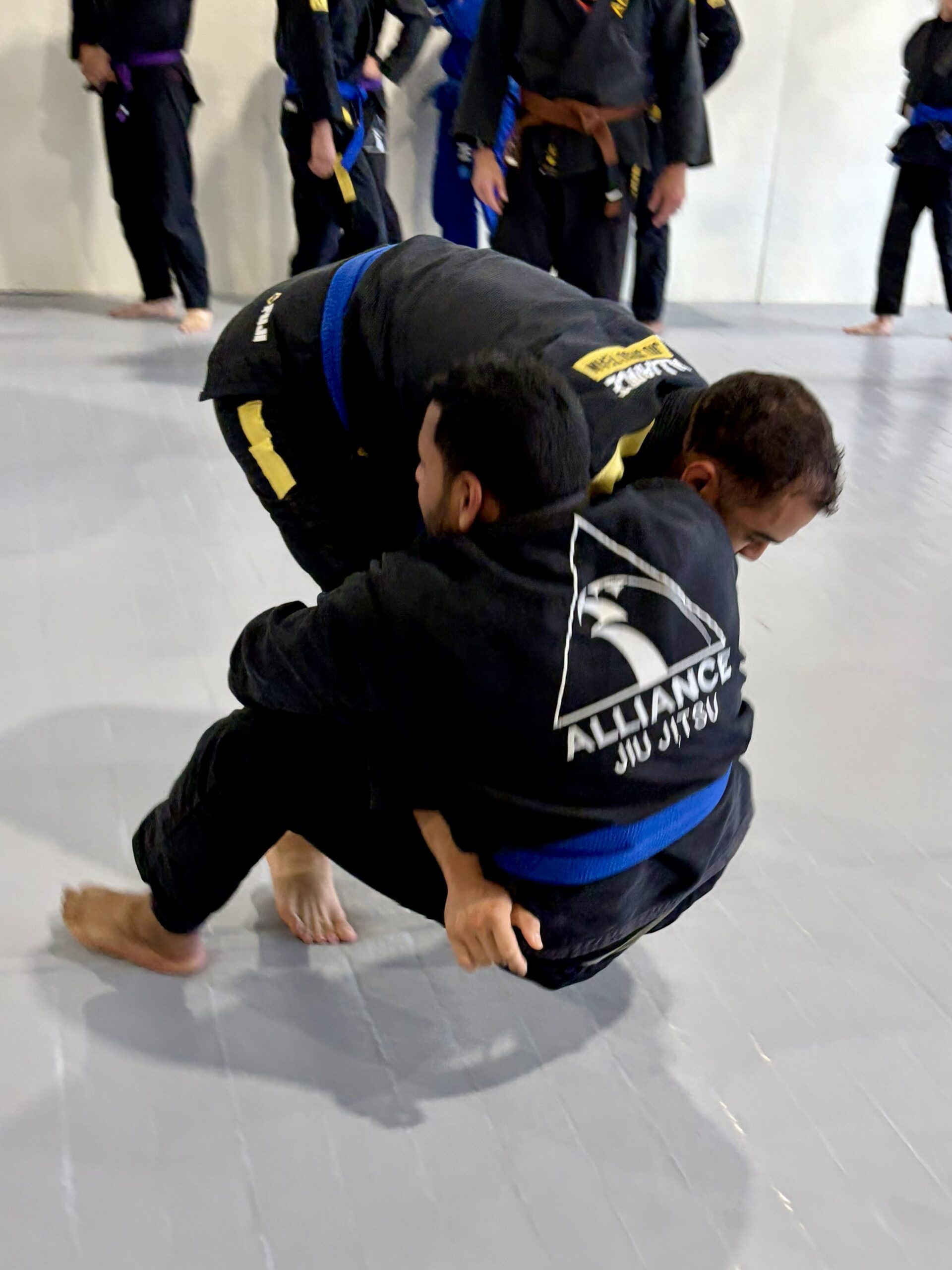 Young man in a black gi controls a kneeling partner from the back during a round