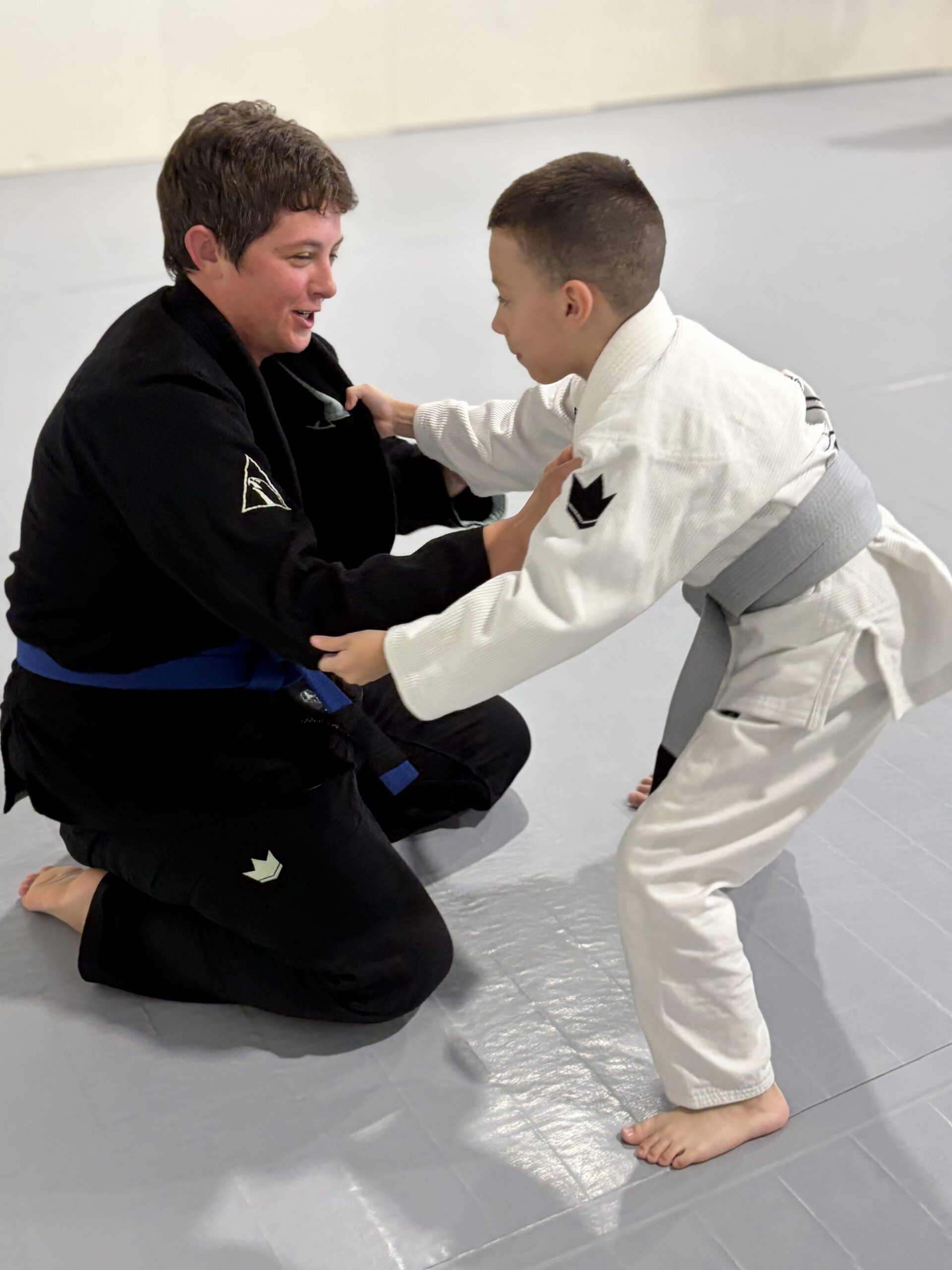 Woman in a black gi with a new blue belt kneels smiling with a small boy
