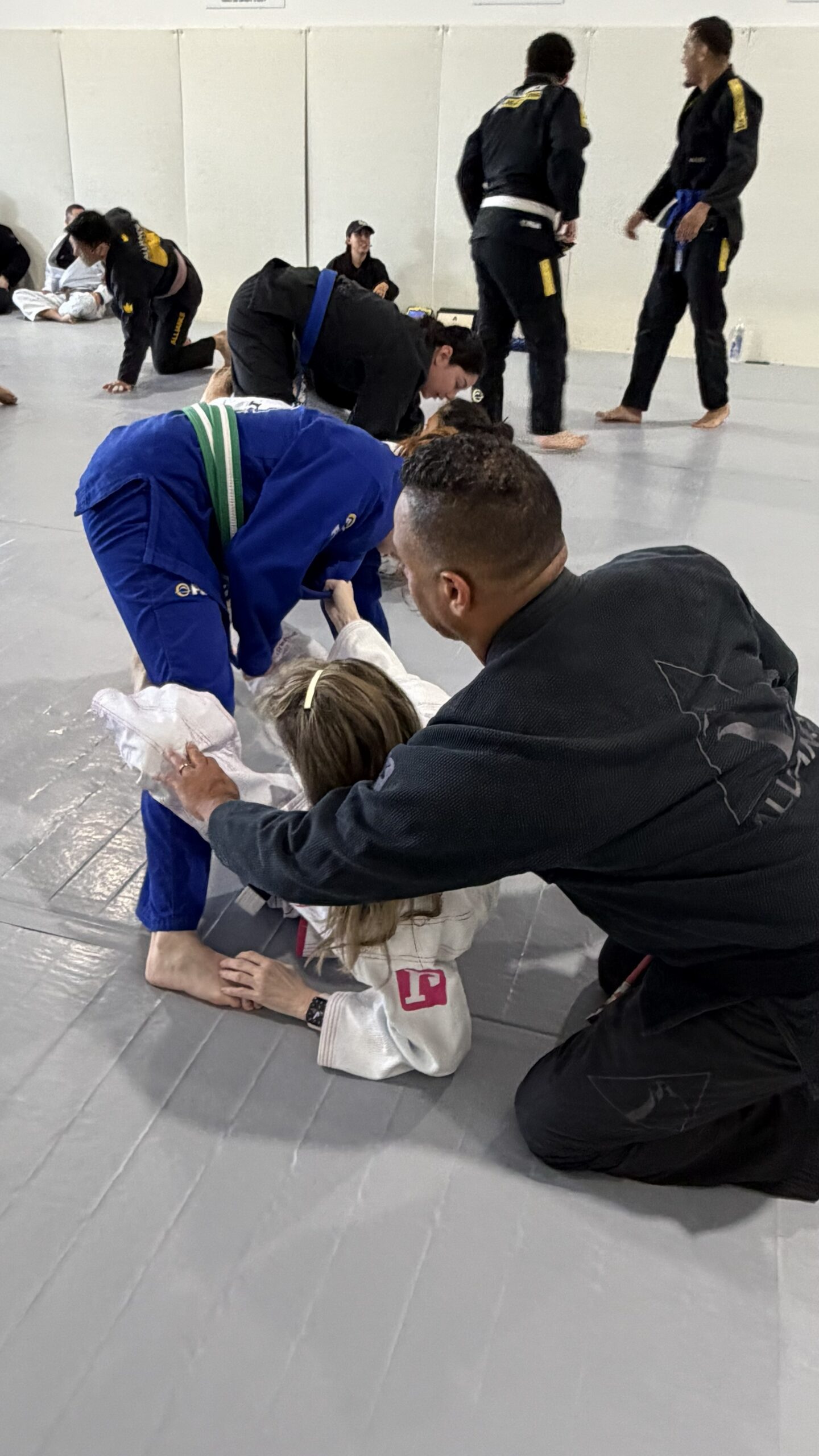 Coach in a black gi helps a young student on a busy academy mat