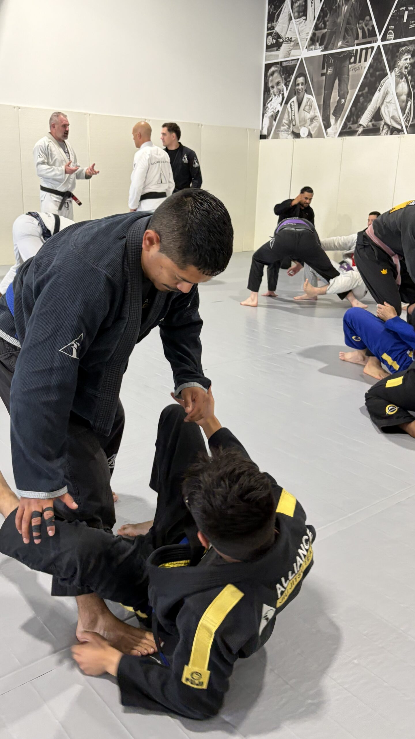 Man in a black gi grips the collar of a kneeling teammate during drilling