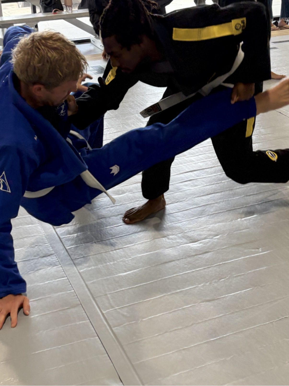 Black belt in a black gi shoots a low single-leg takedown on a partner in blue