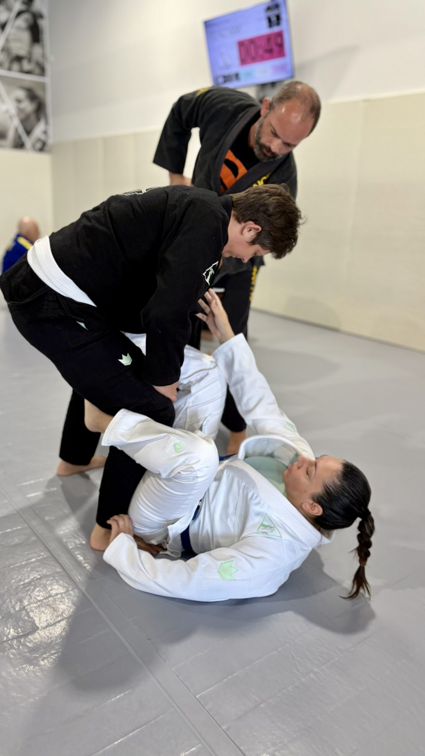 Teen in a black gi passes a woman's guard as a coach watches nearby