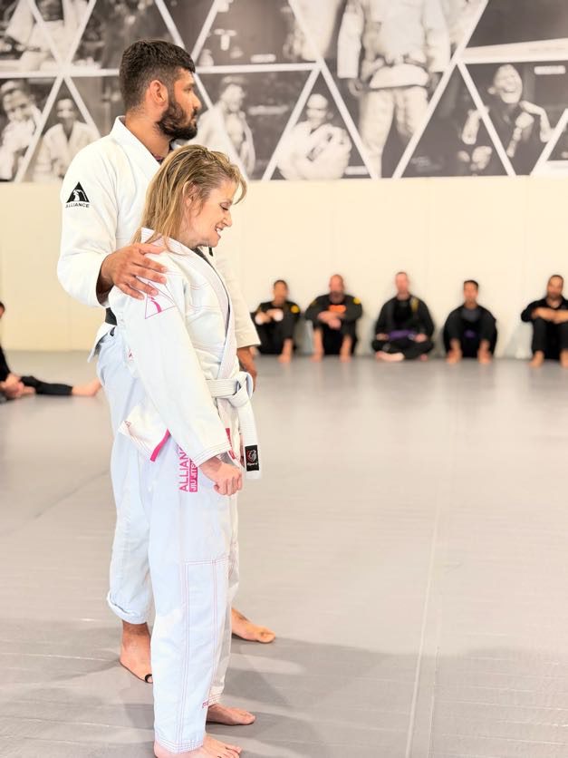 Coach stands behind a smiling student in a white gi to tie her new belt