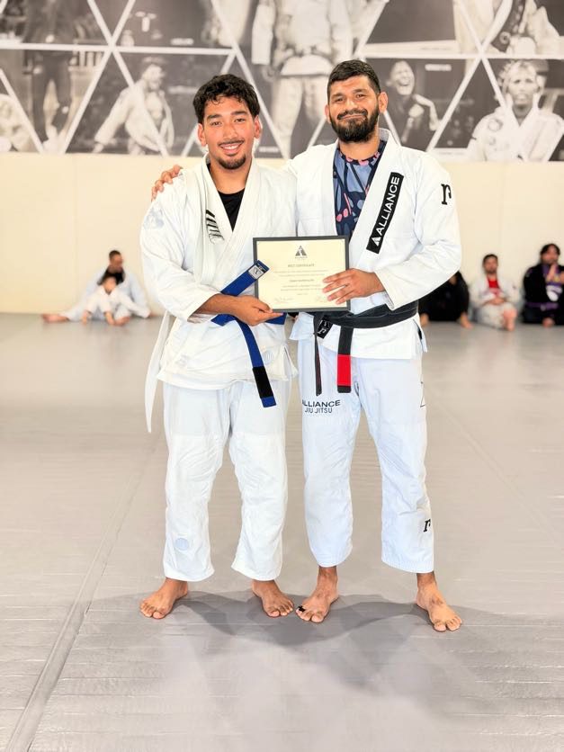 Man in a white gi holds his certificate and new blue belt beside his coach