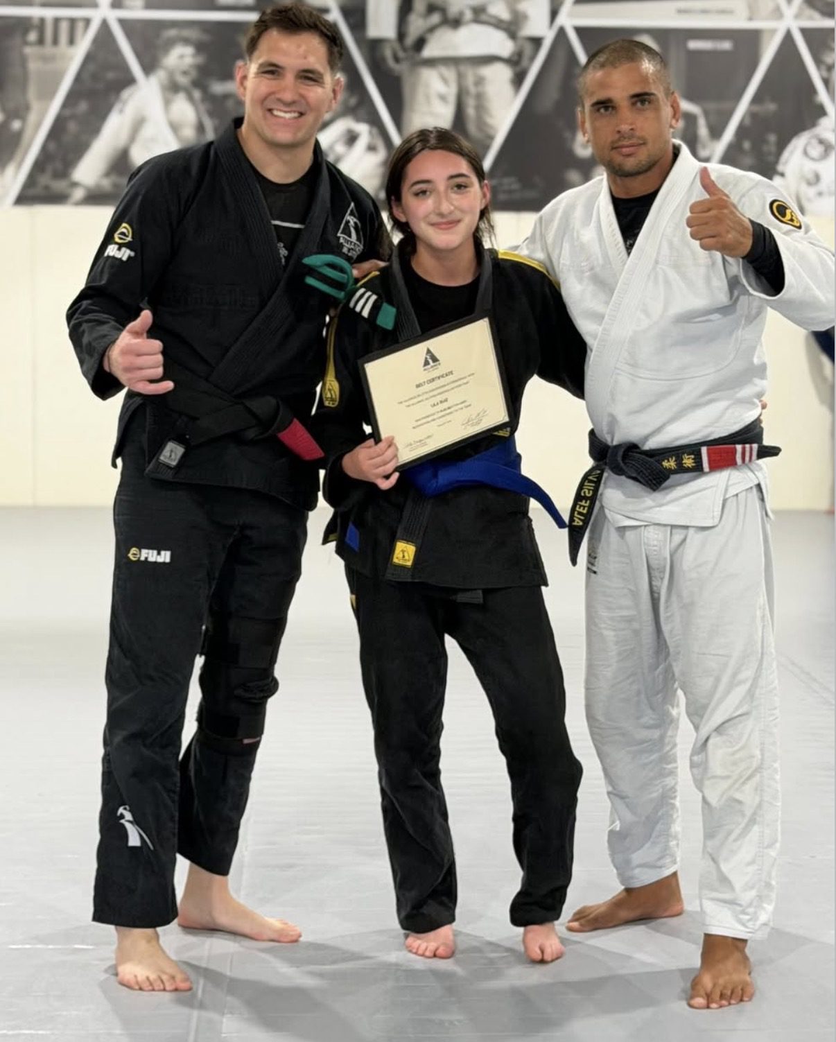 Lila Ruiz in a black gi holds her certificate between two Alliance coaches