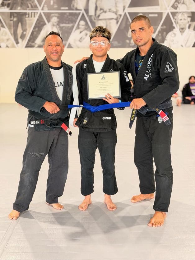 Man in a black gi holds a certificate as coaches present his new blue belt
