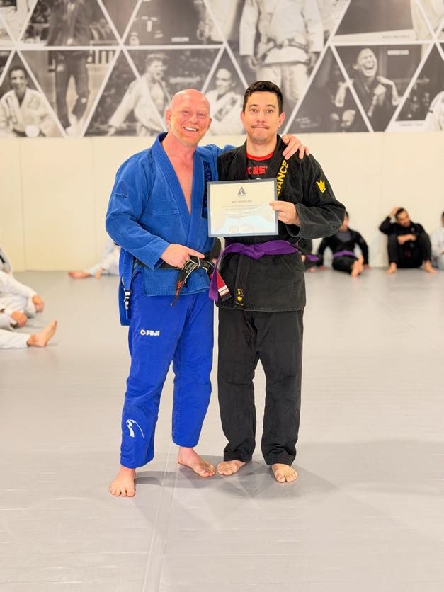 Scott Japalucci in a black gi holds his purple belt certificate beside a coach