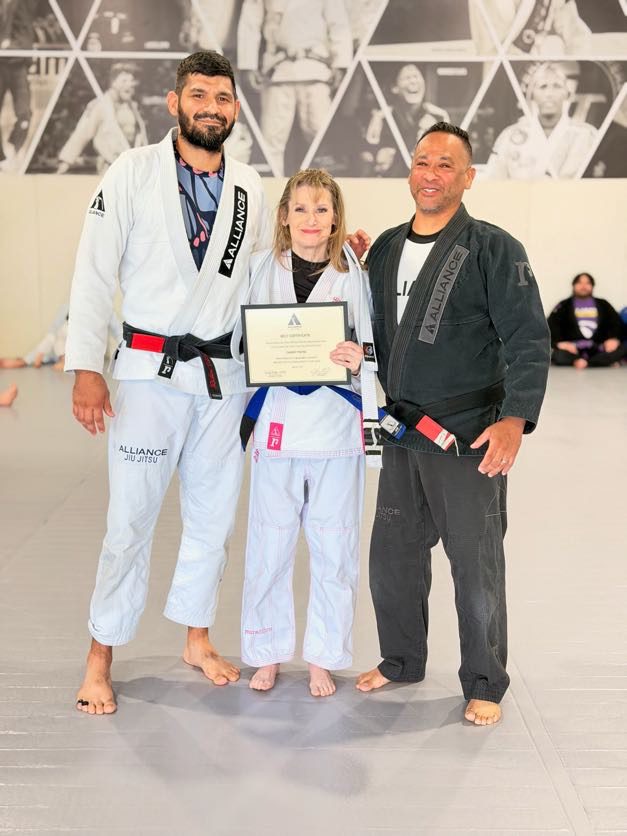 Tammy Payne holds her certificate in a white gi beside two Alliance black belt coaches