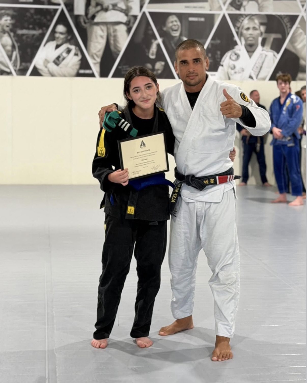 Teen in a black gi holds her new blue belt and certificate beside a coach