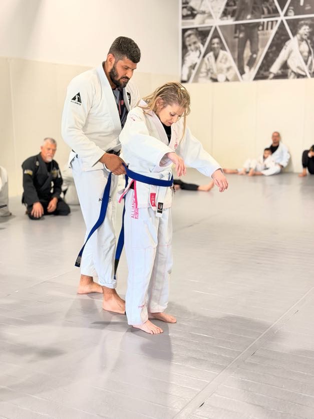 Coach in a white gi ties a blue belt around an adult student during the ceremony