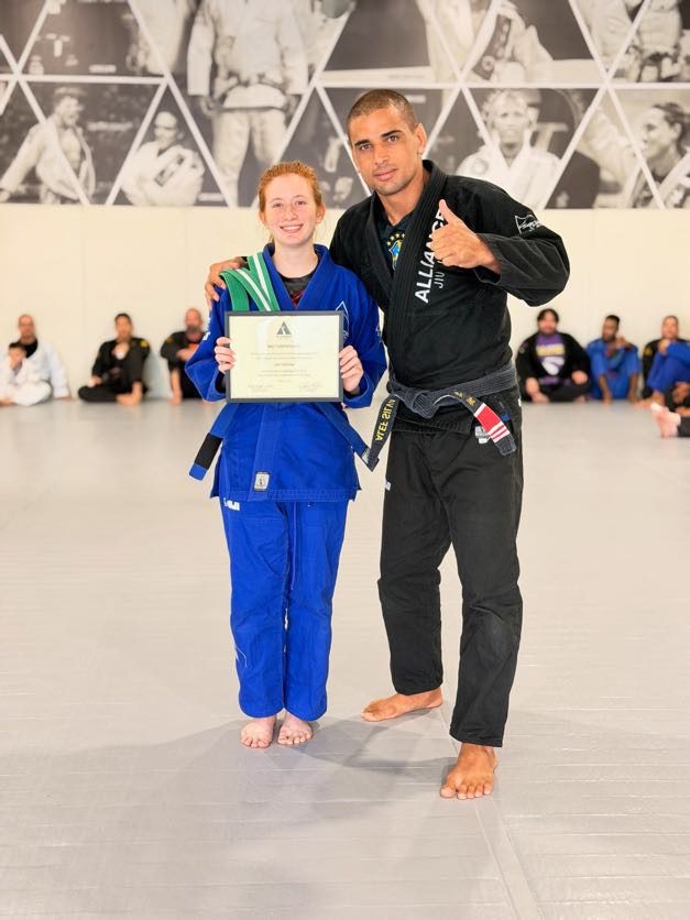 Woman in a blue gi holds her certificate beside a thumbs-up Alliance coach