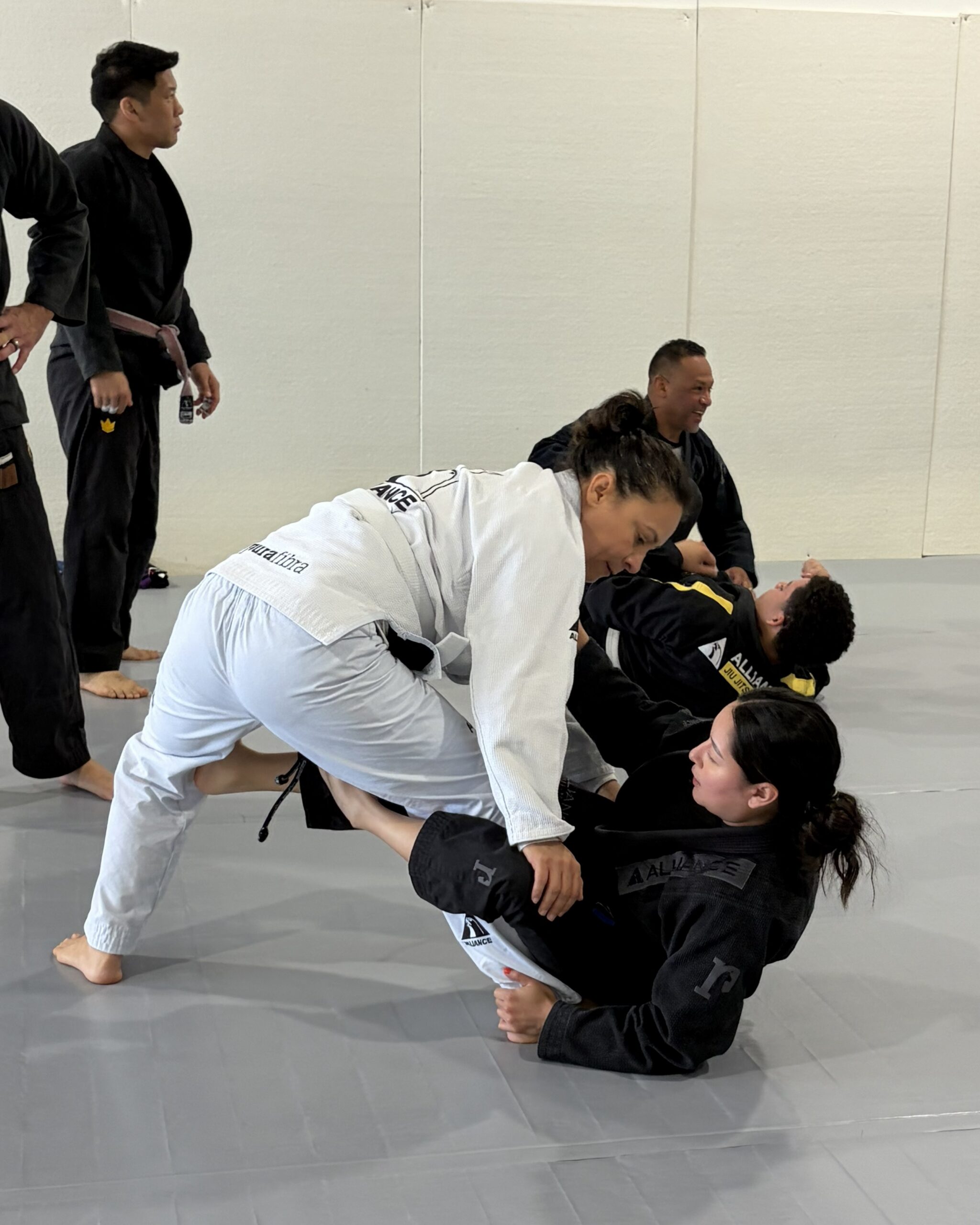 Woman in a white gi passes the guard of two partners in black gis during training