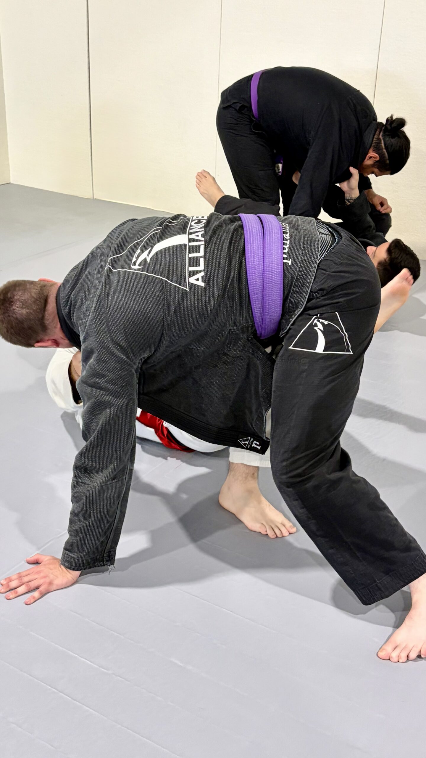 Woman in a black gi and purple belt sprawls on top of a partner on all fours