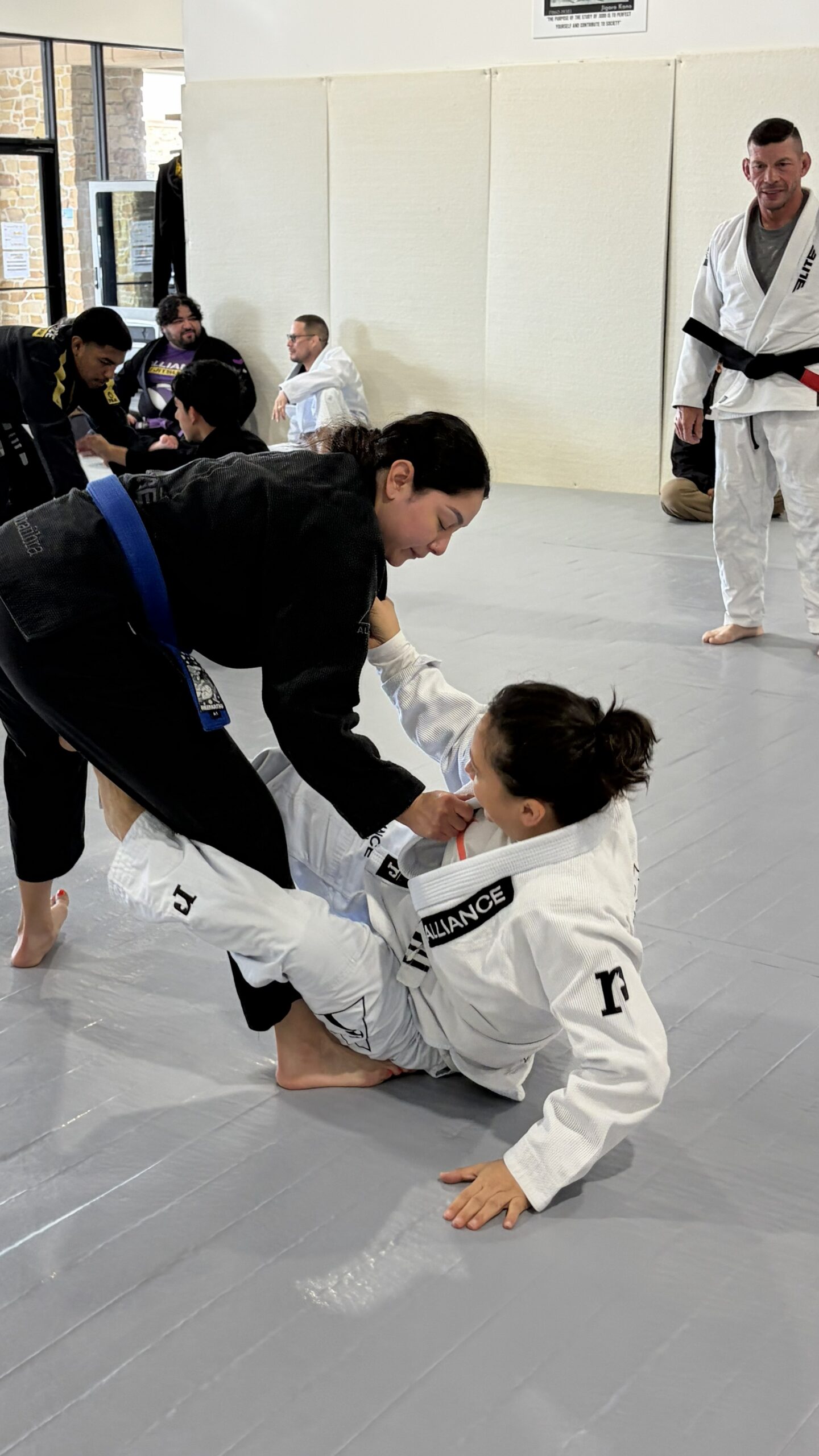 Woman in a black gi and blue belt controls a kneeling partner in a white gi