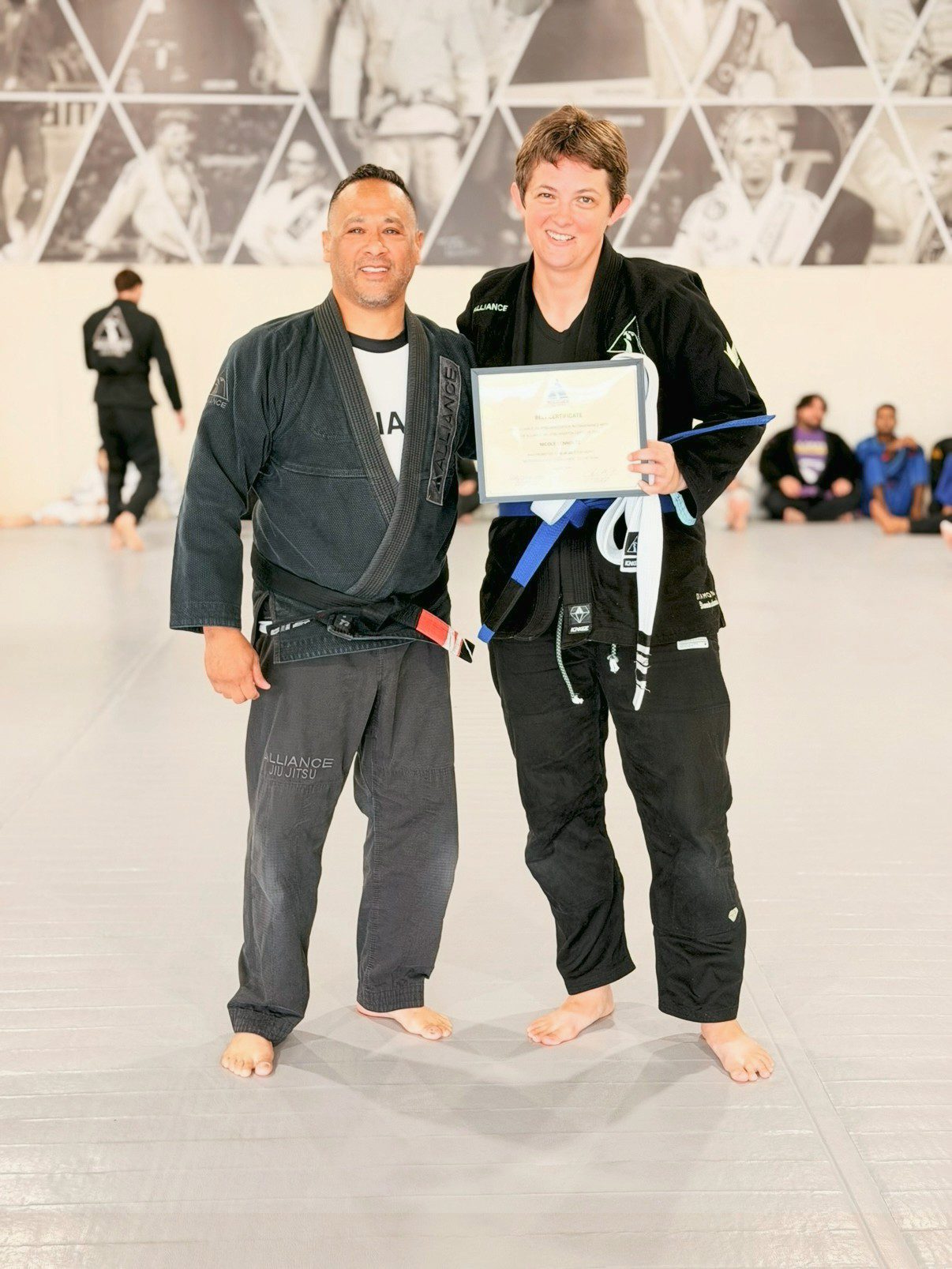 Coach holds a certificate beside a woman in a black gi with her new blue belt