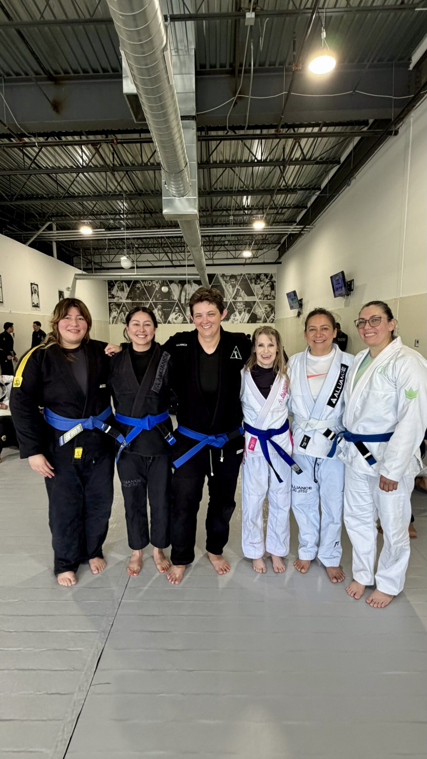 Six women in mixed gis stand arm-in-arm smiling after their Alliance belt promotions