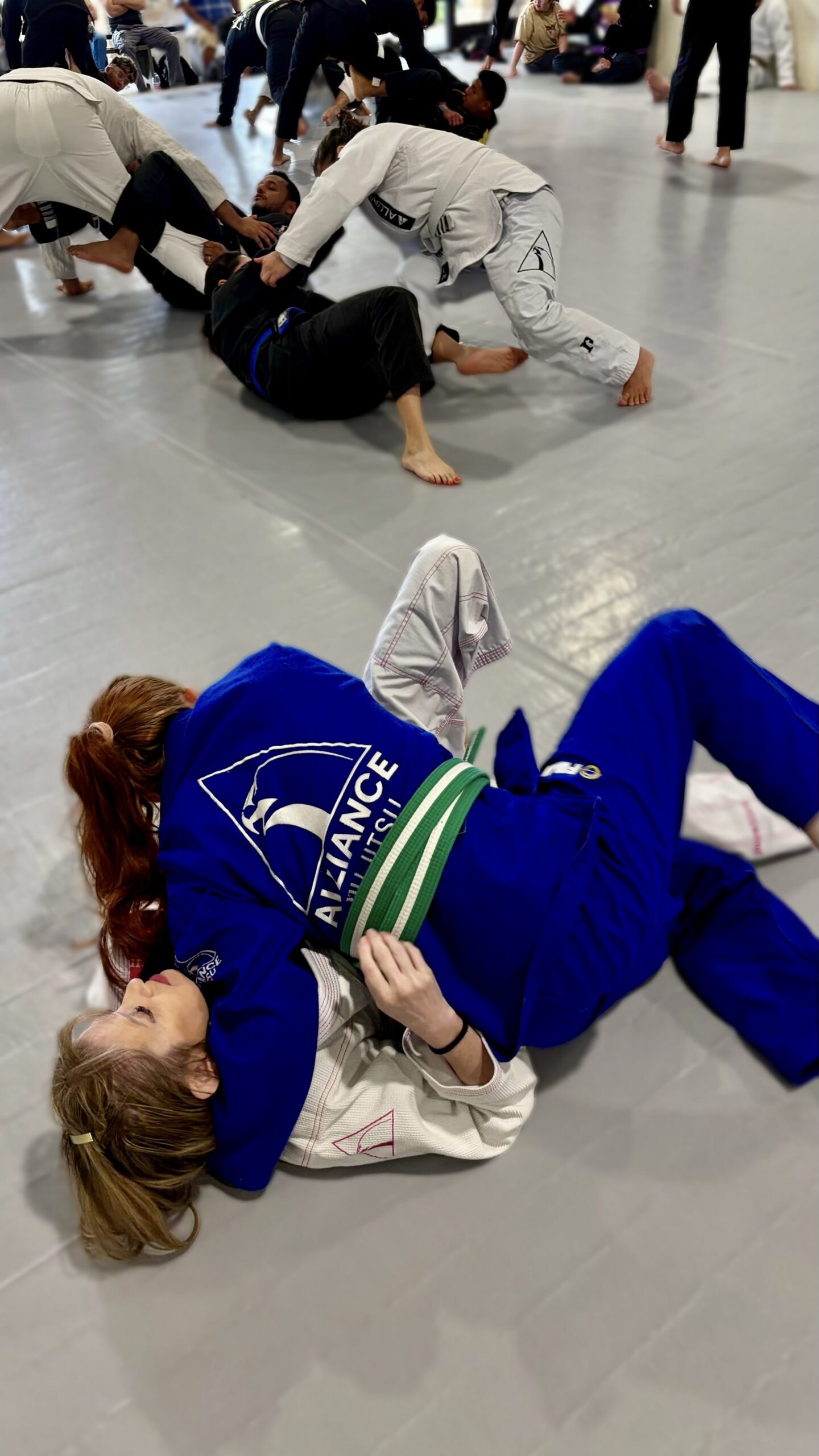 Woman in a blue gi grapples a partner from her back during open mat sparring